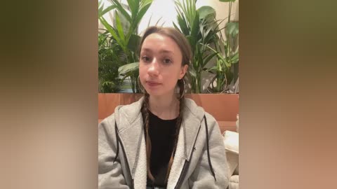 A video of a young woman with light skin and brown hair, wearing a gray cardigan over a black shirt, sitting in a room with lush green potted plants in the background.