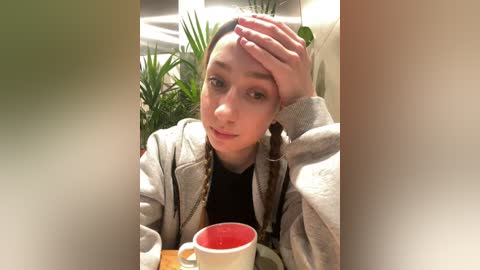 Video of a young woman with light skin, long brown hair, and a grey jacket, holding her forehead while seated at a table, with a red mug and green plant in the background.