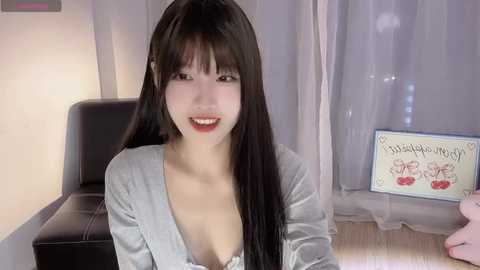 Media: Video of an East Asian woman with long black hair, light skin, and a slim physique, wearing a low-cut gray blouse, smiling in a room with light-colored curtains and a black chair.