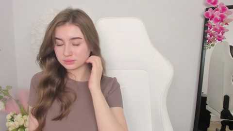 Media: A young Caucasian woman with long brown hair, wearing a short-sleeved brown top, sits in a white gaming chair, adjusting her hair. The background features a white wall and pink floral arrangements.