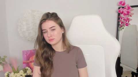 Media: Video of a young Caucasian woman with long brown hair, wearing a grey t-shirt, seated in a white gaming chair in a minimalist room with floral accents.