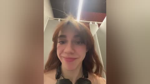 Media: Video of a young woman with long, straight red hair, fair skin, and light makeup, wearing black headphones, smiling in a dimly lit changing room with a red curtain and fluorescent lights.