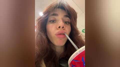 Media: A close-up video of a young woman with wavy brown hair, full lips, and a silver straw in a red can, taken indoors, blurry background.