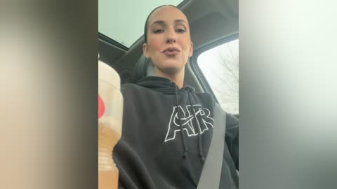 Media: A candid video of a light-skinned woman with a sleek ponytail, wearing a black hoodie with white lettering, sipping from a white and red Starbucks cup while driving.