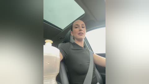 Media: Video of a woman with a slender build and light skin tone, wearing a dark gray shirt and seatbelt, driving a car with a beige steering wheel and window.