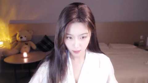 Media: Video of an East Asian woman with long, straight black hair, light skin, wearing a white robe, sitting on a bed with a beige headboard, lit by a bedside lamp.