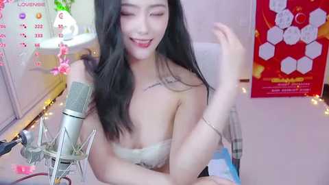 Media: A video of an East Asian woman with long black hair, wearing a strapless, off-white lace bra, sitting at a desk with a microphone, calendar, and bee-themed poster.