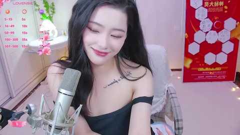 Media: Video of an East Asian woman with long black hair, wearing a black off-shoulder top, smiling while recording herself on a microphone in a pink-themed room with posters and flowers.