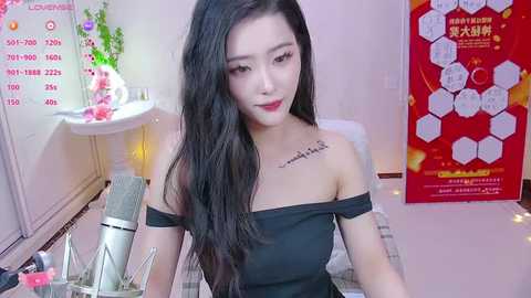 Media: Video of an East Asian woman with long black hair, wearing a black off-shoulder dress, sitting at a desk with a microphone and a calendar in the background.