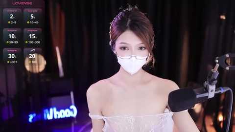 Media: Video of a young woman with light skin, brown hair, and glasses, wearing a white face mask, off-shoulder white lace dress, microphone stand, dimly lit background, digital on-screen stats.