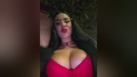 Media: A video of a woman with large breasts in a red bra, wearing glasses and a black jacket, lying on a dark surface.