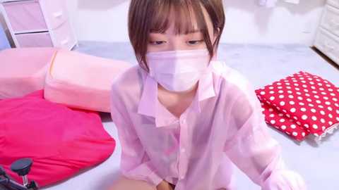 Media: A video of a young Asian woman with a surgical mask, wearing a sheer pink blouse, sitting on a light blue floor with pink and polka-dotted pillows in a minimalistic room.