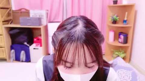 Media: Video of an Asian woman with long, dark hair, wearing a blue school uniform, gagged with white tape, standing in a tidy, pink-themed room with shelves and wicker baskets.