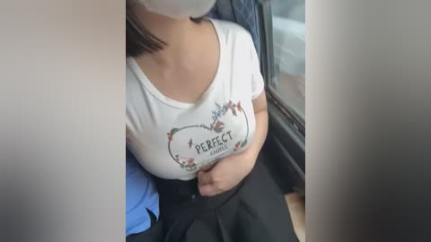 Media: Video of a woman sitting in a car, wearing a white t-shirt with \"Perfect\" text, black skirt, and a face mask.