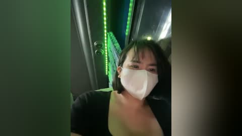 Media: Video of a young Asian woman with short black hair, wearing a black top and white surgical mask, standing in a dimly lit, neon-lit room with green LED strip lights on the walls.