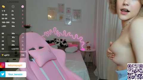 Media: A video of a topless woman with light skin and short blonde hair, standing in a pink gaming chair in a cozy bedroom. The room features a bed, white curtains, and a pink neon sign.