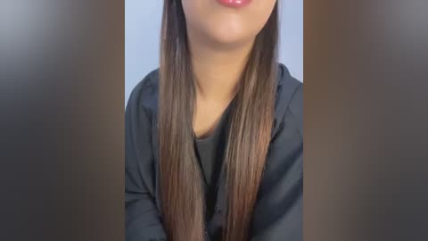 Media: Video of a woman with long, straight, brown hair parted down the middle, wearing a black top. The background is a blurred, neutral tone.