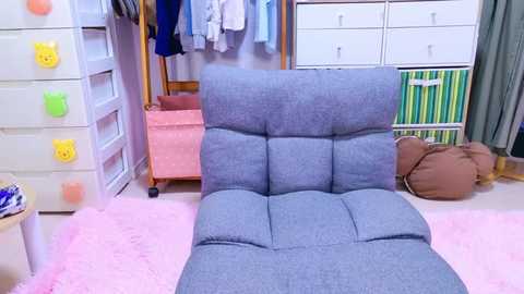 Media: Video of a cozy, pastel-toned bedroom with a plush, light gray, tufted chair on a pink shaggy rug. The background features a white dresser, a clothes rack, and storage bins.