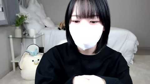 Media: A video of an East Asian person with straight black hair, wearing a black mask, sitting on a plush carpet, holding a plush toy in a modern, minimally decorated room.