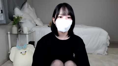 Media: A video of a young Asian woman with short black hair, wearing a black T-shirt and white mask, sitting on a plush bear in a white, minimalist bedroom.