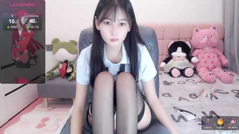 Media: Video of a young Asian woman with long black hair, sitting on a gray chair in a room with plush toys and a pink bed.