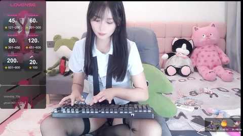 Media: A video of an Asian woman with long black hair, wearing a white shirt, black tie, and thigh-high stockings, typing on a keyboard in a room with pink cushions and stuffed animals.