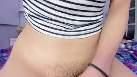 Media: Video of a woman with light skin, wearing a black-and-white striped crop top, exposing her pubic hair. Background shows a purple wall and a bed with colorful bedding.