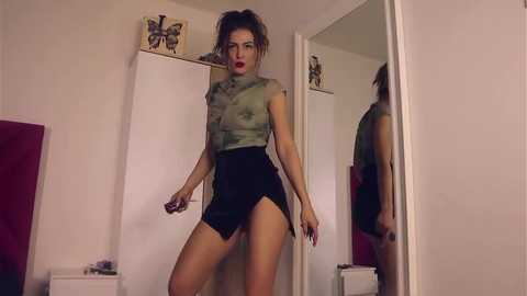 Media: Video of a slender, light-skinned woman with dark hair in a ponytail, wearing a green camisole and black high-waisted shorts, standing in a dimly lit bedroom with white walls and a red bed.