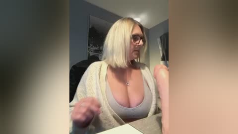 Media: Video of a middle-aged Caucasian woman with short blonde hair, glasses, wearing a cream-colored knit cardigan over a low-cut gray top, seated at a desk.