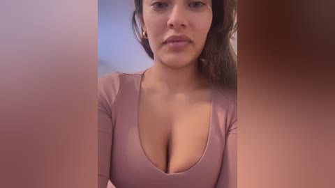 Media: A close-up video of a woman with medium brown skin, long brown hair, and a low-cut, pink long-sleeve top, revealing ample cleavage, against a blurred, reddish-brown background.