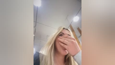 Media: Video of a young woman with long blonde hair, covering her face with one hand, standing in a modern, well-lit room with white walls and ceiling.