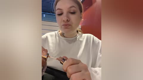 Media: A video of a young woman with light skin, brown hair, and a nose ring, wearing a white shirt, applying makeup with a brush and lipstick in a bathroom with a red towel and closed window.