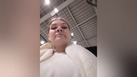 Media: A video of a young woman with fair skin and blonde hair, wearing a white bathrobe, standing in an industrial warehouse with exposed ceiling beams and fluorescent lights.