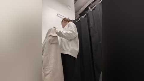 Media: Video of a man in a white lab coat and black pants, hanging a beige coat in a stark, minimalist changing room with black curtains.