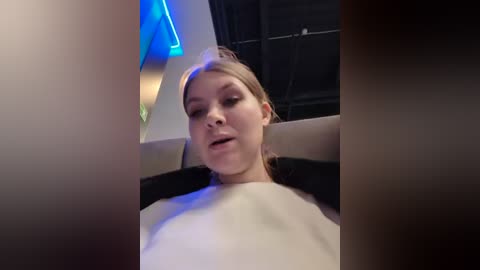 Media: Video of a fair-skinned woman with blonde hair, wearing a white top, reclining in a modern, dimly lit room with a blue neon sign in the background.