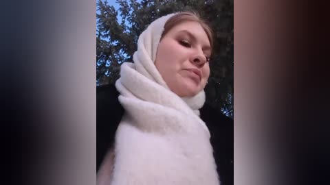 Media: Video of a young Caucasian woman with light skin, wearing a white woolen scarf and a black jacket, standing outdoors against a backdrop of leafy trees. She has blonde hair partially covered by the scarf, and her expression is serene.