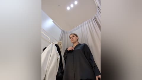 Media: Video of a young woman in a fitting room, wearing a black blouse, standing in front of a white curtain, with a white coat and black jacket hanging nearby.