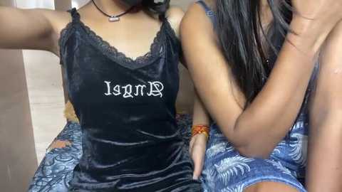 Media: Video of two women, one wearing a black velvet camisole with \"Isis\" embroidered, the other a blue floral dress, sitting closely together, with a blurred background.