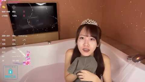 Media: Video of an Asian woman with long black hair, wearing a silver tiara, sitting in a pink bathtub with a green towel, surrounded by water droplets, in a beige bathroom.