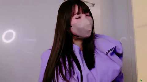 Media: Video of a young Asian woman with long black hair, wearing a lavender bathrobe and a white face mask, standing in a white-tiled bathroom, eyes closed, appearing relaxed.