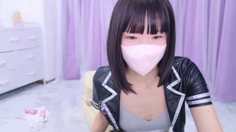 Media: Video of an Asian woman with straight black hair and blunt bangs, wearing a white mask and a black leather jacket over a gray bralette, sitting on a white floor with light purple curtains and white drawers in the background.