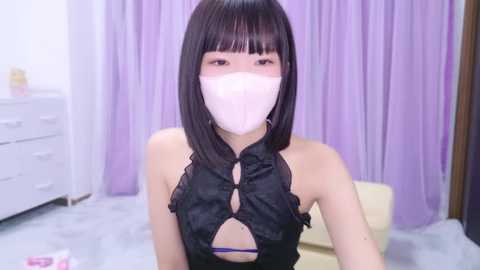 Media: A video of an Asian woman with straight black hair, wearing a black halter top, white face mask, and purple underwear, sitting in a room with lavender curtains and white furniture.