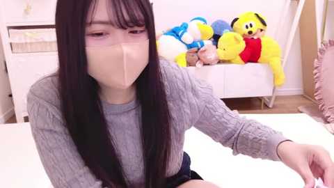 Media: Video of an Asian woman with long black hair, wearing a gray sweater, beige tape over her mouth, seated indoors with stuffed toys in the background.