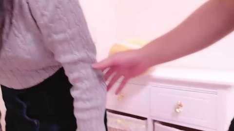 Media: Video of a person in a light gray, textured sweater, with a hand touching their arm, standing in front of a white dresser with drawers, in a bright, clean bathroom.