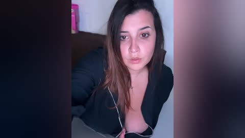 Media: Video of a young, light-skinned Latina woman with long, dark hair, wearing a black, unbuttoned jacket, puckering her lips, in a dimly lit room with a pink bottle on a nightstand.