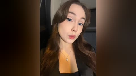 Media: Video of a young woman with long brown hair, light skin, and blue eyes, wearing a black top and gold necklace. She puckers her lips and stares directly at the camera.