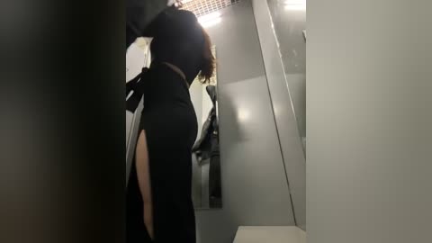 Media: A video of a woman with light skin and shoulder-length brown hair, wearing a black dress, standing in a dimly lit, narrow hallway with gray walls and a metal door.