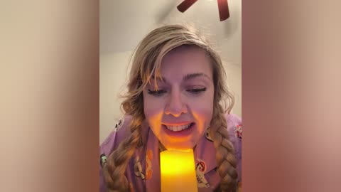 Media: A video of a blonde woman with wavy hair, wearing a purple shirt with colorful patterns, smiling with closed eyes, holding a lit yellow candle in front of her face, set against a soft, blurred background.