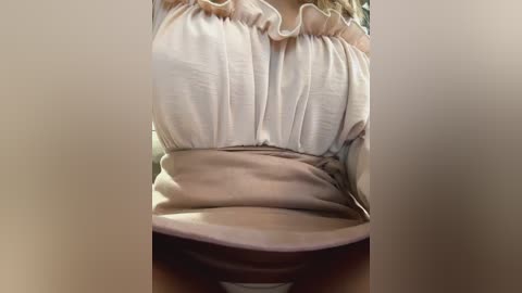 Media: Video of a woman's torso, wearing a cream-colored blouse with ruffled sleeves and a beige belt cinched at the waist, partially revealing her brown skirt.