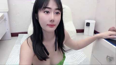 Media: A video of an Asian woman with long black hair, fair skin, and small breasts, wearing a green dress, sitting on a white chair in a minimalistic, modern room with white walls and a striped rug.